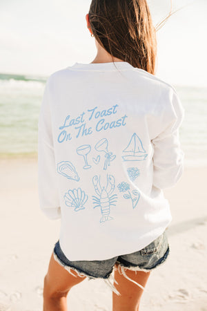 A girl at the beach wears a white sweatshirt with a blue Last Toast On The Coast design printed on the back