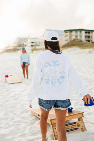 A girl plays cornhole in a white sweatshirt with a blue Last Toast On The Coast design printed on the back.