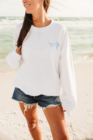 A girl on the beach wears a white sweatshirt with a blue bow design and "Kendra's Bach"