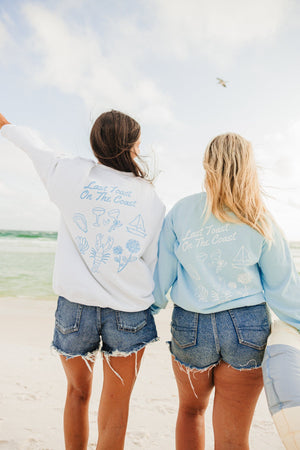 Two girls at the beach wear a white and a blue sweatshirt with a matching Last Toast On The Coast design printed on the back