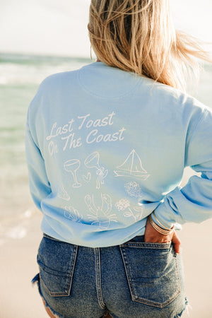 A girl at the beach wears a blue sweatshirt with a white Last Toast On The Coast design printed on the back