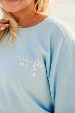 A girl wears a blue sweatshirt that is customized with a white bow design that says "Kendra's Bach"