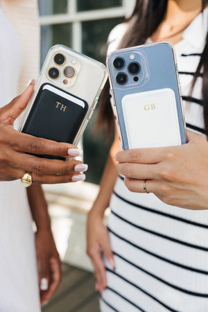 Close-up of two women holding iPhones with transparent cases, each adorned with a monogrammed card holder. The left holder is black with a 'TH' monogram in gold, and the right one is white with a 'GB' monogram also in gold. This detailed view highlights the elegant personalization on a practical accessory, set against the blurred background of a casual outdoor setting.