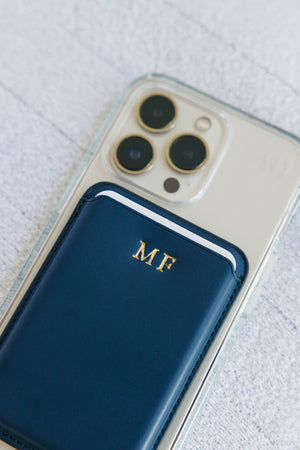 Two individuals holding up their smartphones, each encased in a transparent cover with a personalized card holder. The left phone features a black card holder monogrammed with 'TH' in gold, while the right one sports a white card holder with a gold 'GB' monogram. This image captures the stylish functionality of the accessories in a casual outdoor setting, emphasizing the personalization of everyday tech.