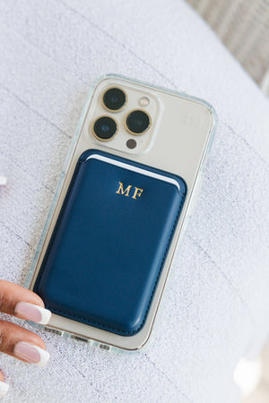 Two women holding smartphones with transparent cases, each showcasing a personalized card holder. The left phone features a black card holder with a gold 'TH' monogram, while the right phone has a white card holder with a gold 'GB' monogram. The image captures the stylish and functional aspect of the accessories, with a subtle urban backdrop adding to the modern aesthetic.