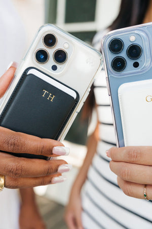 A smartphone with a transparent case featuring a navy blue card holder with a personalized gold 'MF' monogram. The phone and card holder are positioned on a textured white fabric surface, emphasizing the sleek design and sophisticated personal touch of the accessory.