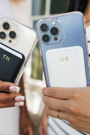 An assortment of smartphones with personalized card holders displayed on a pink and white checkered blanket. The collection includes various colors and monograms: black with 'AL', white with 'KD', navy with 'J', tan with 'MD', white with 'GB', black with 'TH', and navy with 'MF'. This vibrant display showcases the customizable options available for a stylish and practical phone accessory.