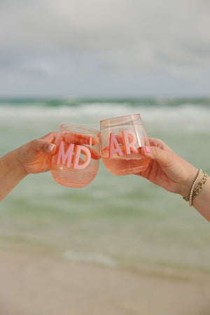 Two people at the beach cheers with pink wine glasses with pink monograms printed on them.