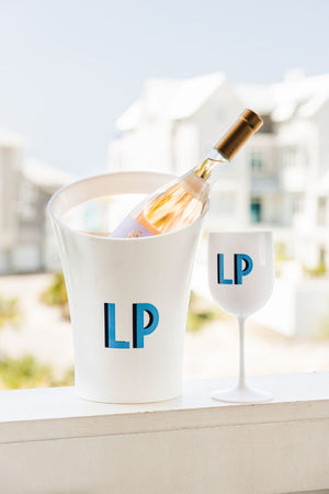 A white ice bucket with a bottle of wine is placed next to a white wine glass with a matching blue monogram.