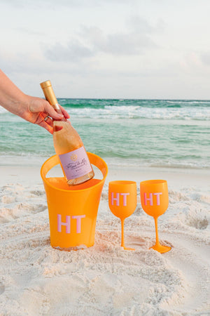 A person grabs a bottle of wine out of an orange ice bucket to be poured into two orange wine glasses with matching pink monograms.