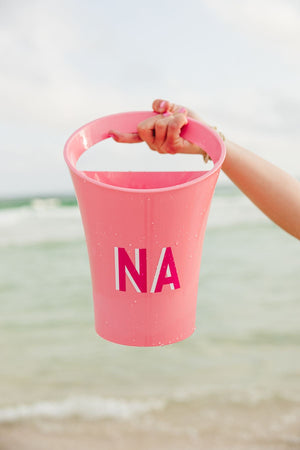 A person holds up a pink monogrammed ice bucket by the water at the beach