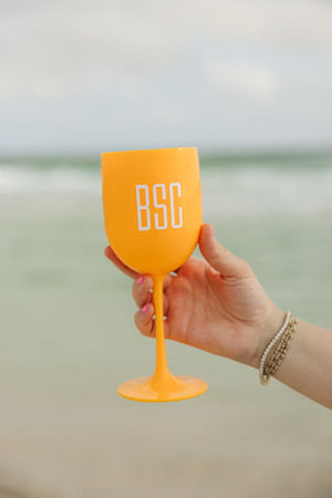 A person at the beach holds up an orange wine glass with a white monogram.