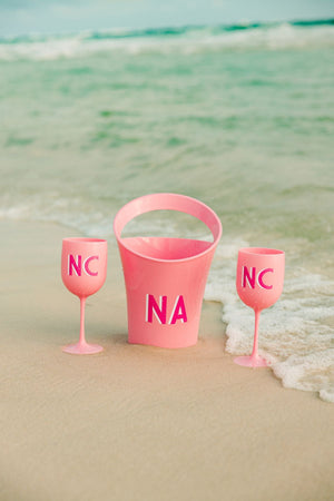 A pink ice bucket and two pink wine glasses are placed on the beach with matching pink monograms