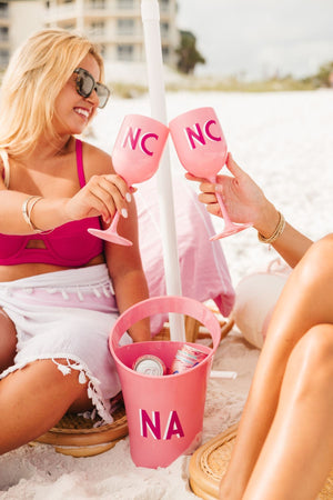 Two people at the beach cheers with their pink wine glasses next to their pink ice bucket.