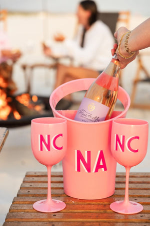 A person pulls a bottle of wine out of a pink ice bucket which has been placed by two pink wine glasses with matching pink monograms