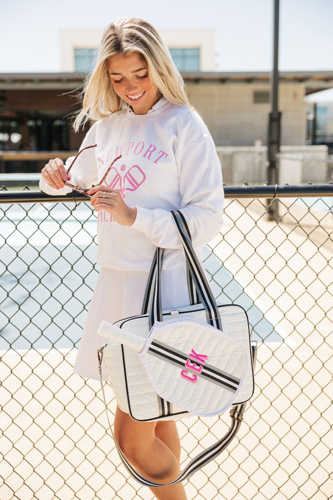 A girl in a white pickleball outfit stand along a fence holding a monogrammed silver and black striped pickleball bag.