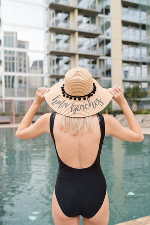 A woman stands by a pool wearing a floppy beach hat with black pom poms and black text that reads "hola beaches"