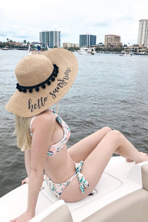 A woman sits by water wearing a floppy beach hat with black pom poms and black text that reads "hello sunshine."