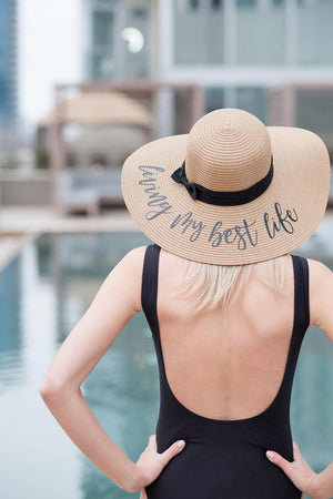 A woman in a black swimsuit shows off her custom beach hat which reads "living my best life."