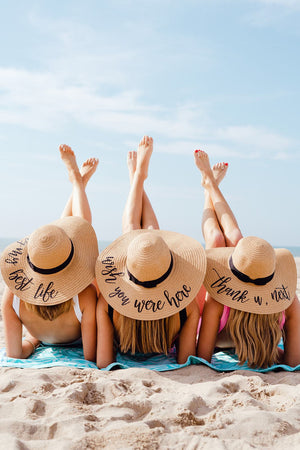 Three women lay on the beach with their feet in the air while they show off their floppy beach hats with customized fun phrased