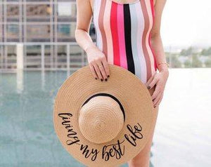 A woman in a stripped swimsuit holds her custom beach hat which reads "living my best life."