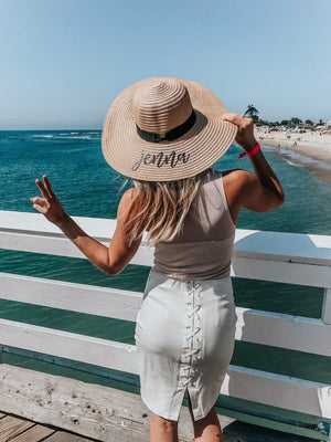 A woman stands on a pier holding up a peace sign and is showing off her floppy hat which is customized with her name