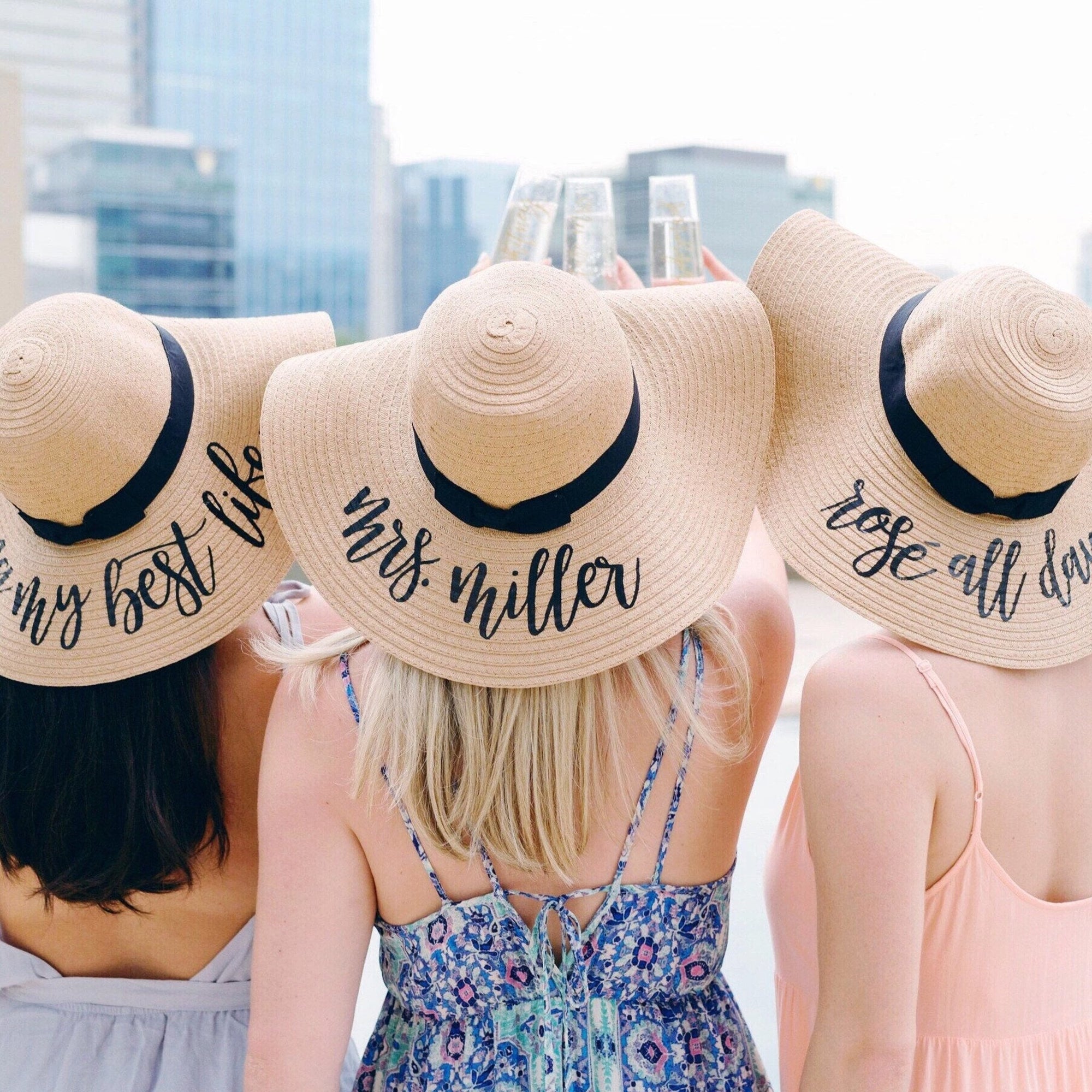 Three women cheers their champagne flutes while wearing customized black ribbon floppy beach hats