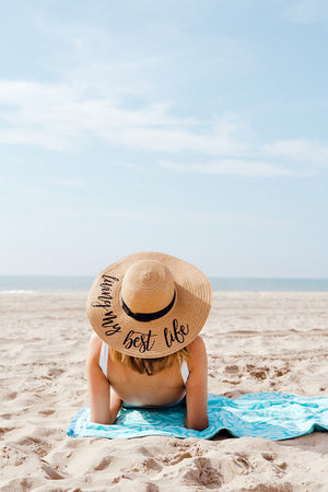 A woman lays on the beach on a blue towel wearing a floppy beach hat customized to read "Living my best life"