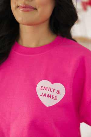 A woman shows off her bright pink sweatshirt personalized with "Emily & James" in a heart.