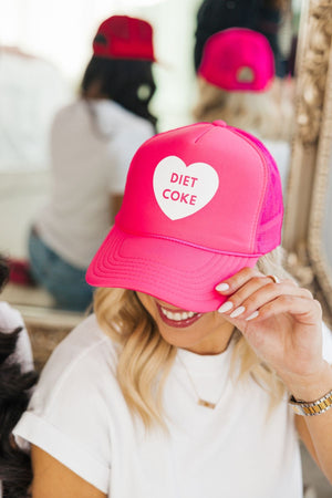 A woman looks down to show off her bright pink trucker hat which reads "Diet Coke" in a heart.