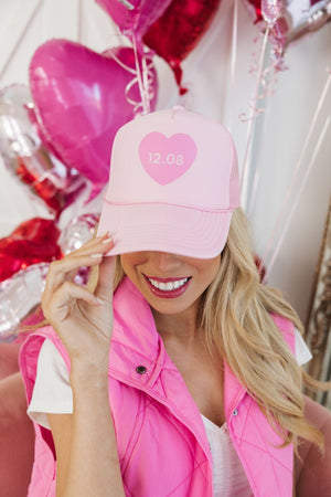 A woman wears a pink trucker hat which is personalized with a date inside the heart design.