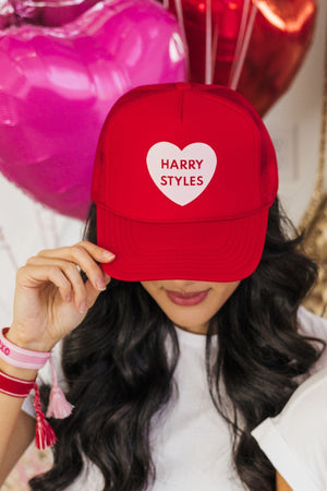 A woman looks down to show off her red trucker hat which reads "Harry Styles" in a pink heart.