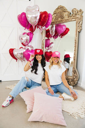 Two women sit showing off their pink and red customized Valentine's Day Hats