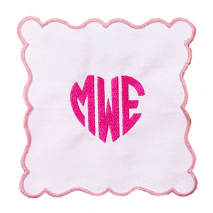 A pink scalloped cocktail napkin is customized with a pink heart monogram