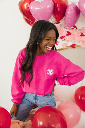 A woman smiles wearing a bright pink sweatshirt embellished with her monogram in a heart