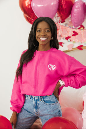 A woman smiles wearing a bright pink sweatshirt embellished with her monogram in a heart