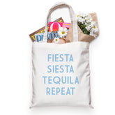 A cotton tote reads "Fiesta Siesta Tequila Repeat" in a light blue font and holds a bouquet of flowers and some magazines.